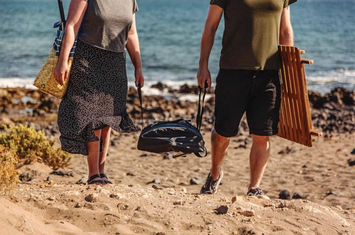 Two people carrying portable grilling tools and accessories on a sandy beach for outdoor living and grilling collections