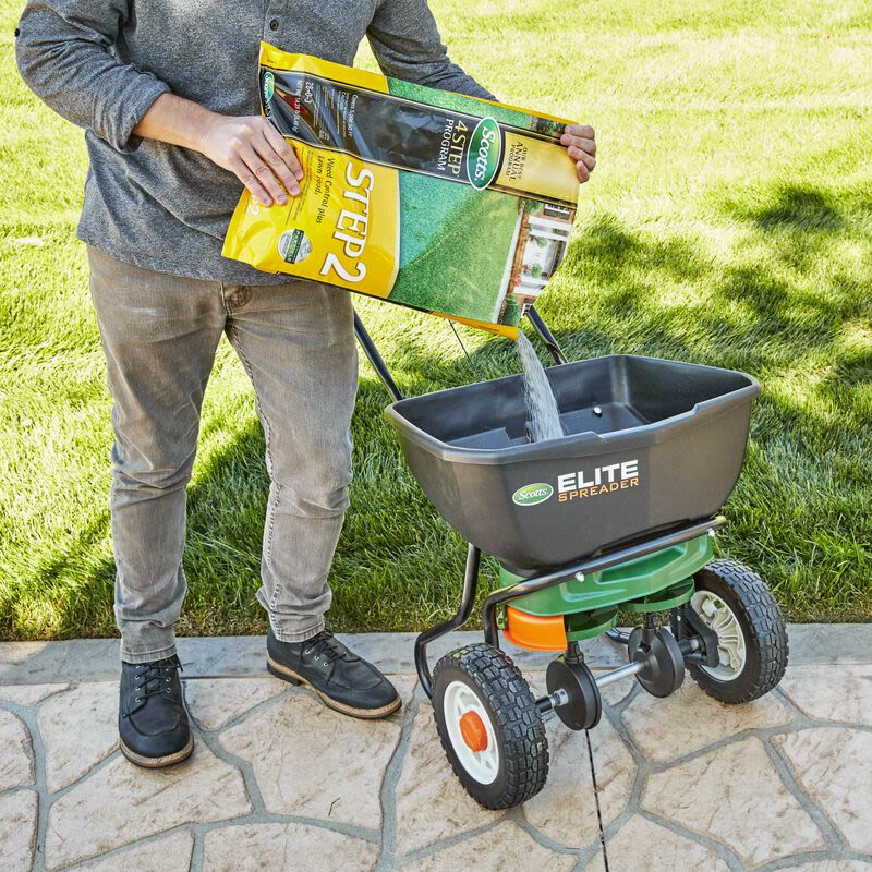 Person pouring weed control and lawn food from a large bag into a wheeled spreader on a stone patio in a lawn care setting