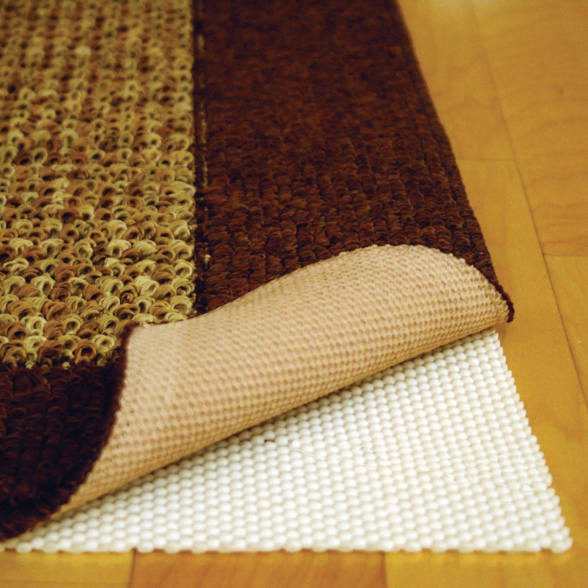 Close-up of a brown and beige area rug corner lifted to reveal a textured nonslip rug pad underneath on hardwood floor