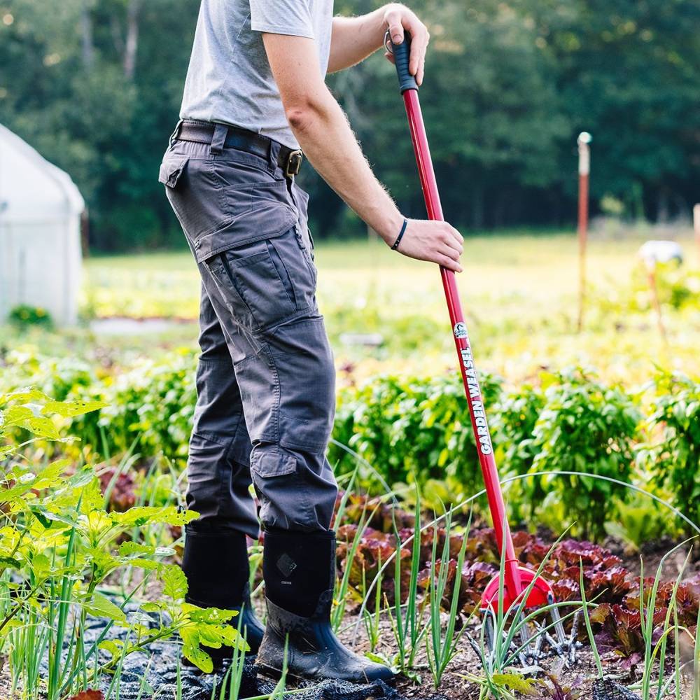 Person using red-handled steel cultivator tilling soil around leafy vegetables in a garden setting