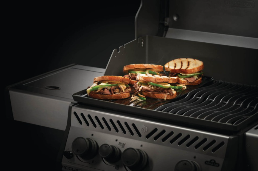 Cast iron reversible griddle on a gas grill cooking toasted sandwiches, ideal for outdoor living and grilling enthusiasts.