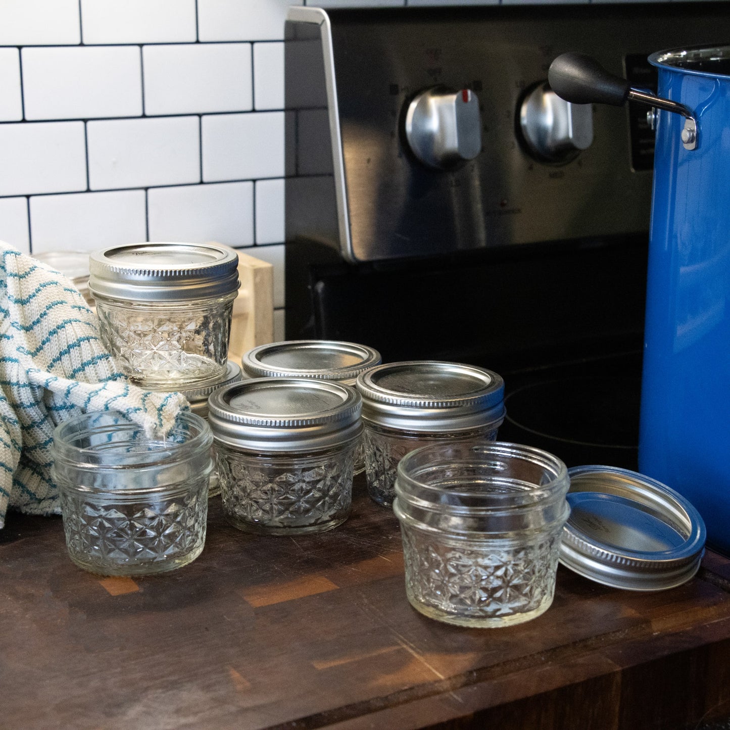 Set of clear 4 oz. glass jars with silver lids on wooden kitchen counter next to stove and blue pot