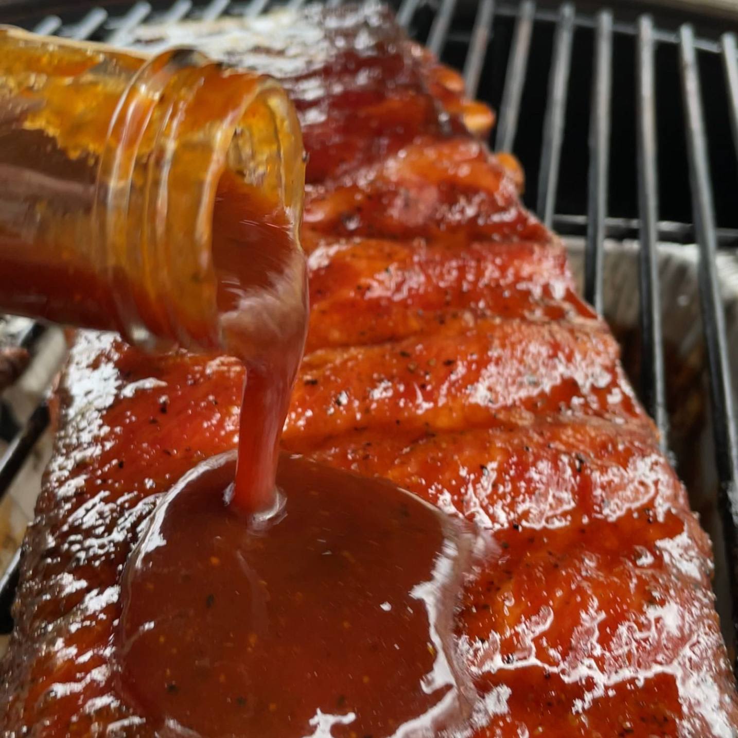 Rich barbecue sauce being poured onto grilled ribs on an outdoor grill in BBQ Sauces & Rubs collection