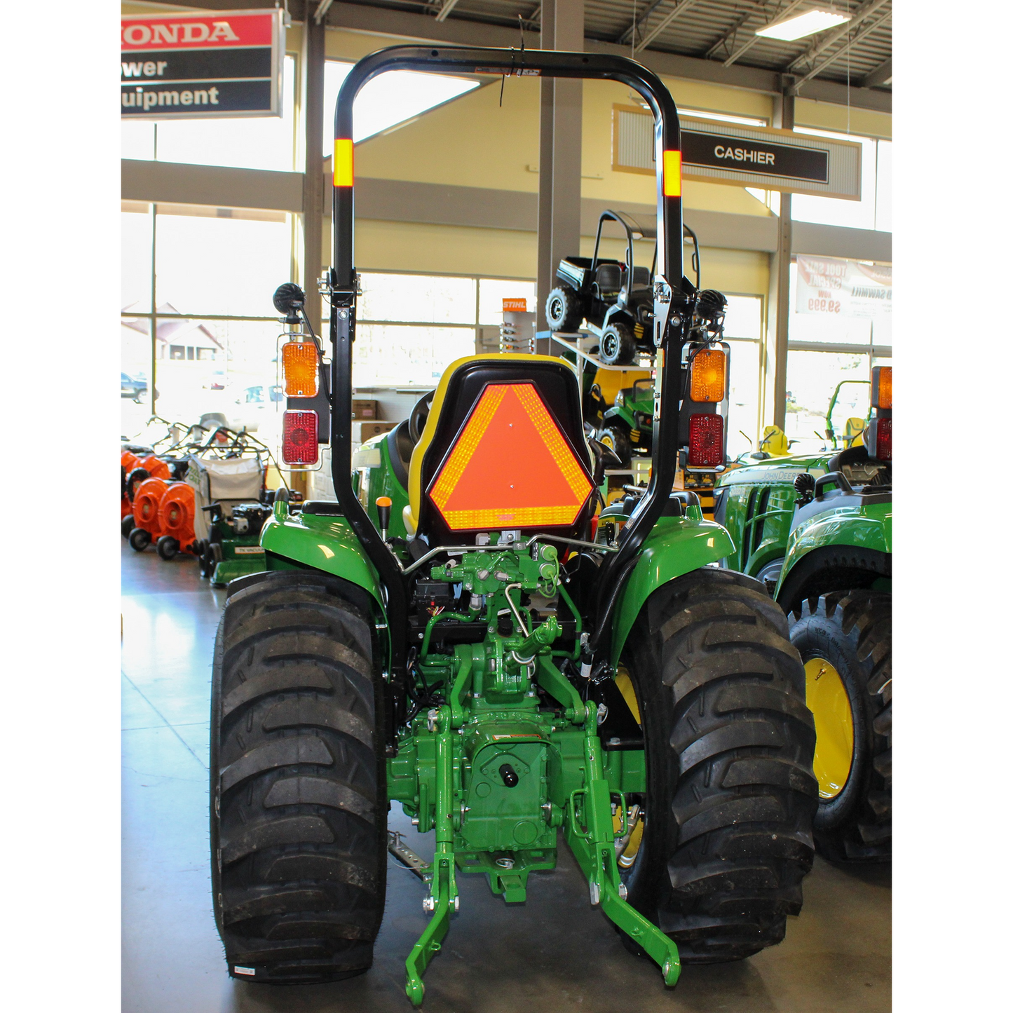 Rear view of a green and yellow John Deere compact utility tractor with large tread tires and safety triangle, displayed indoors among outdoor power equipment and tools collections.