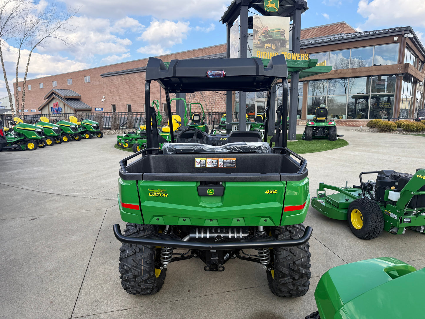 Rear view of a green John Deere mid-size crossover utility vehicle with 4x4 capability, parked outdoors at a dealership among other John Deere equipment and UTVs.