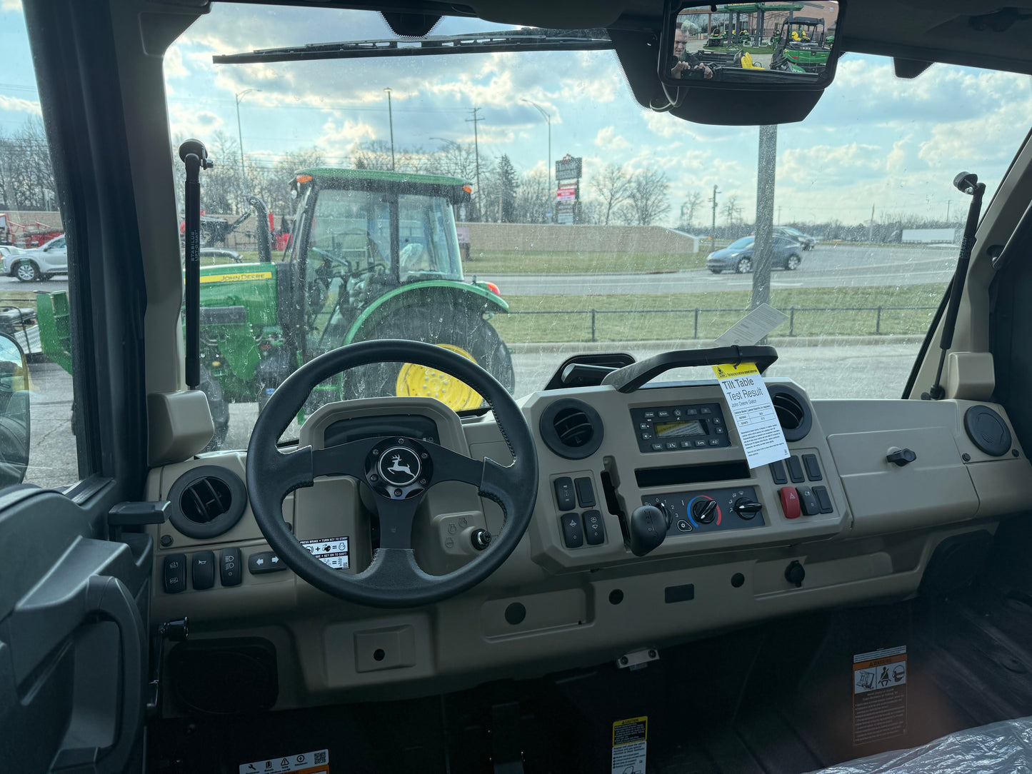 View of the dashboard and steering wheel inside a John Deere JD697731 Custom XUV 845R utility vehicle at an outdoor equipment lot with a tractor visible through the windshield.
