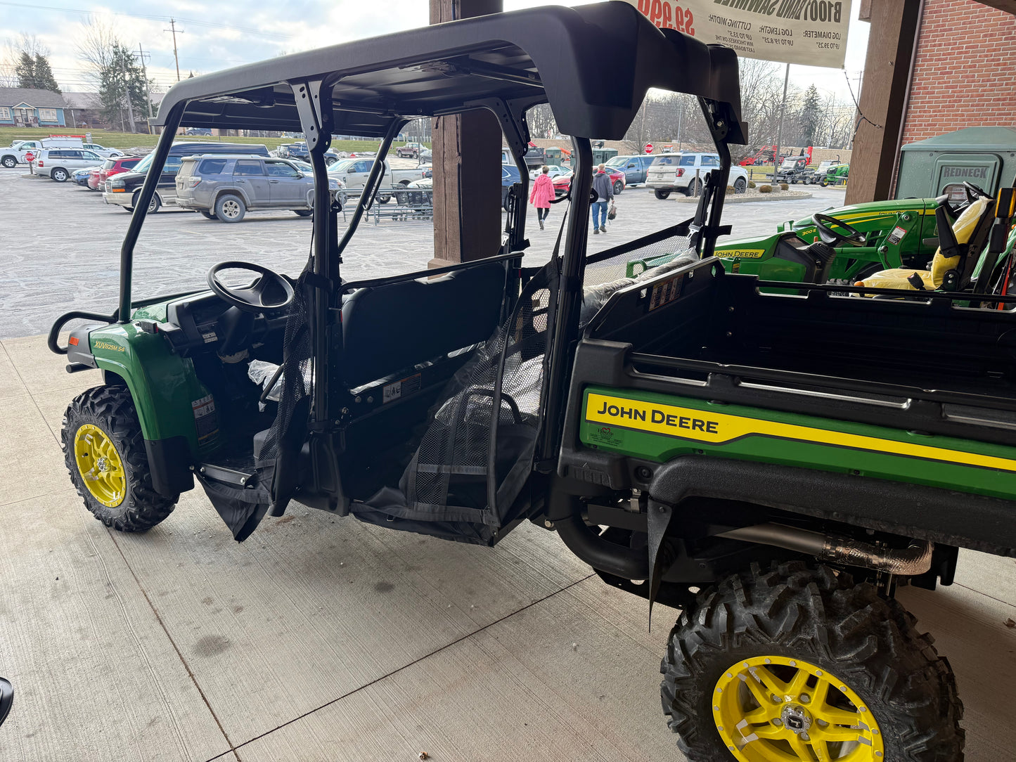 John Deere green and yellow 4-seater crossover utility vehicle with rugged tires and open cabin, parked near equipment at a dealership lot.