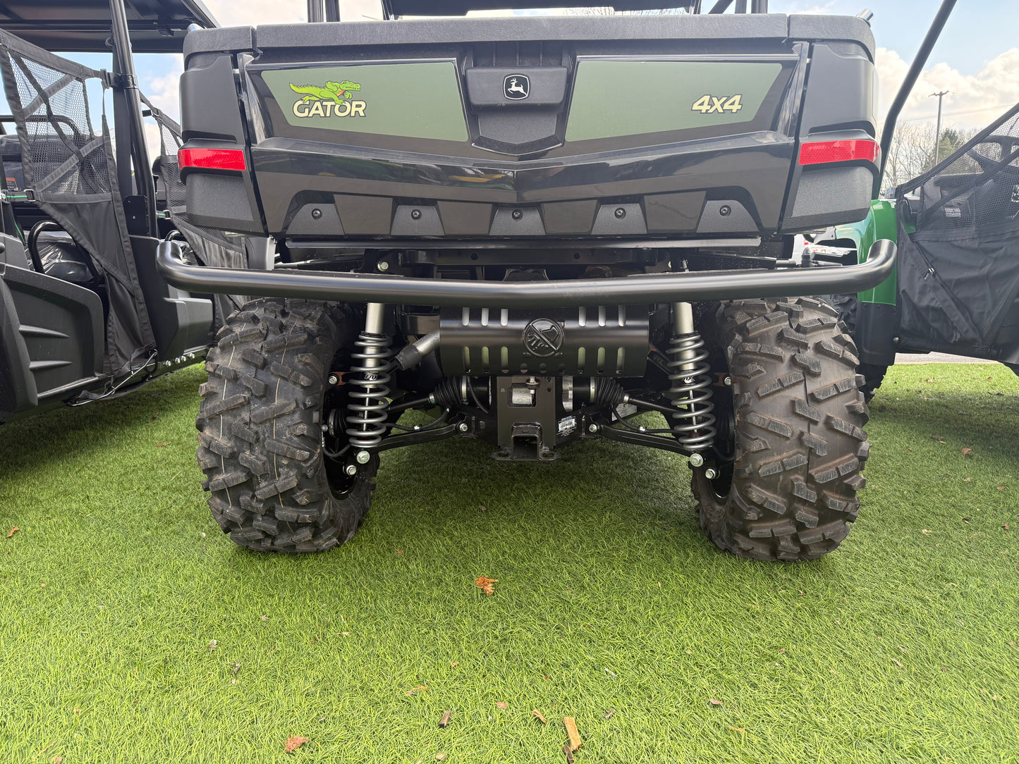 Rear close-up of a green John Deere Gator 4x4 utility vehicle showing suspension, rugged tires, and protective rear bumper on artificial grass at dealership lot