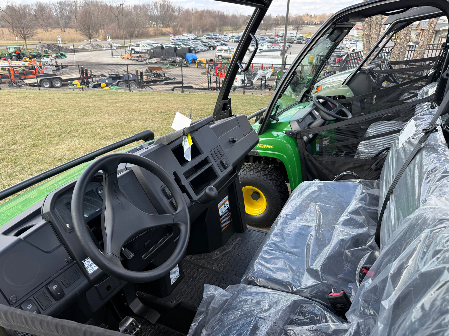 Interior view of a green John Deere XUV utility vehicle featuring steering wheel, dashboard, and protective seat covers at an outdoor equipment dealership lot