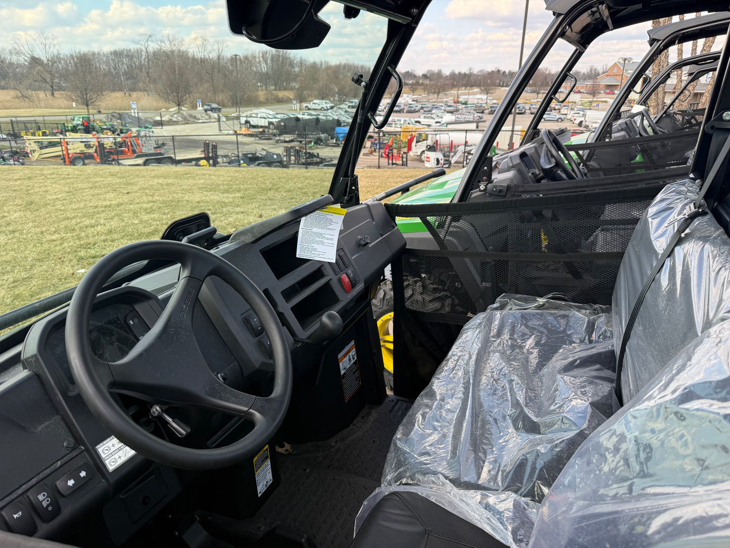 Interior view of John Deere utility vehicle showcasing steering wheel, dashboard, and protective plastic-covered seating at outdoor dealership lot with equipment in background
