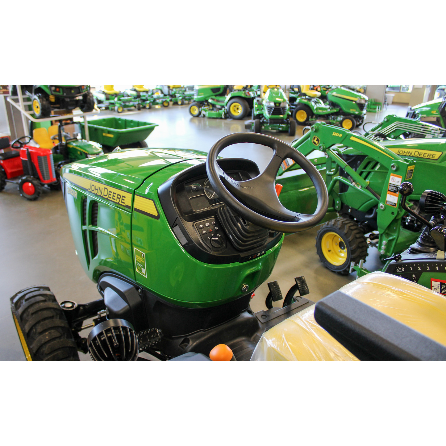 Green John Deere compact utility tractor featuring a detailed dashboard, steering wheel, and yellow seat, displayed in an indoor equipment showroom among outdoor power tools and lawn & garden machinery.
