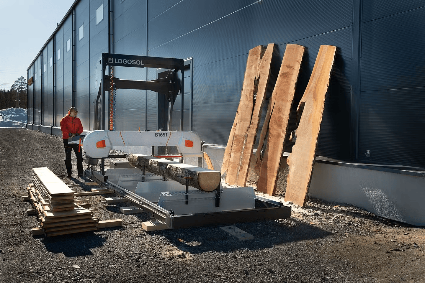 LOGOSOL B1651 band sawmill cutting large log into lumber slabs outdoors near industrial building