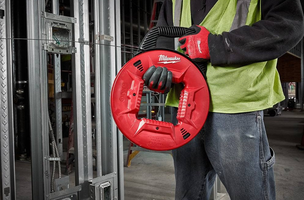 Person wearing work gloves and safety vest holding red steel fish tape tool for wire pulling in construction site