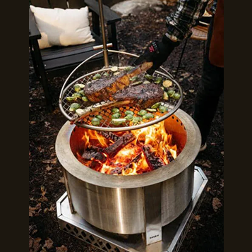 Round metal cooking grate with adjustable pole grilling steaks and vegetables over outdoor fire pit in patio living setting