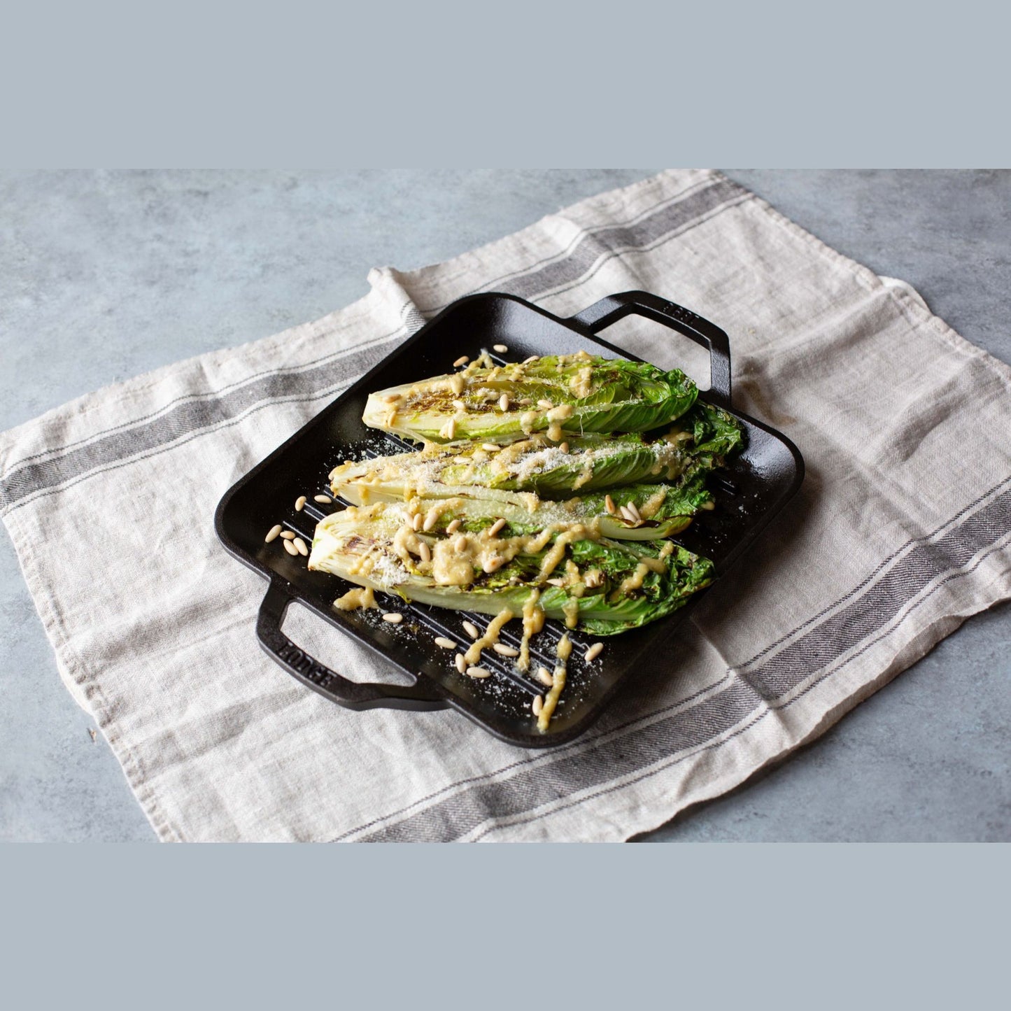 Black cast iron square grill pan with grilled romaine lettuce and creamy dressing on neutral cloth surface