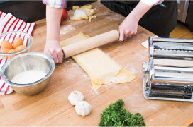 Person rolling fresh pasta dough on wooden countertop with pasta machine and kitchenware tools nearby for homemade ravioli
