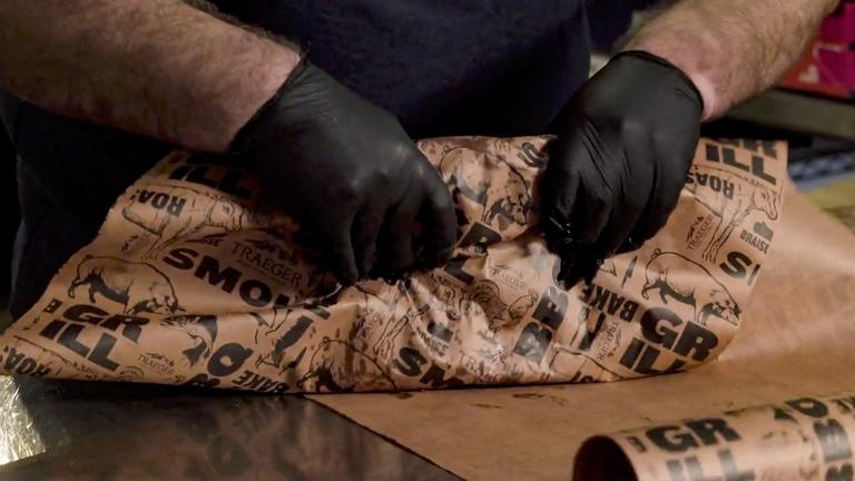 Hands wearing black gloves wrapping food in brown butcher paper with BBQ-themed print for grilling accessories.