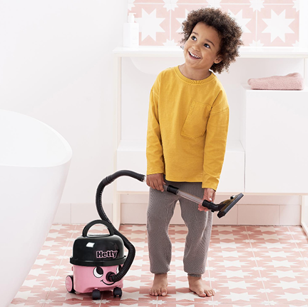 Young child playing with a pink Hetty toy vacuum cleaner on patterned tile floor in bright room