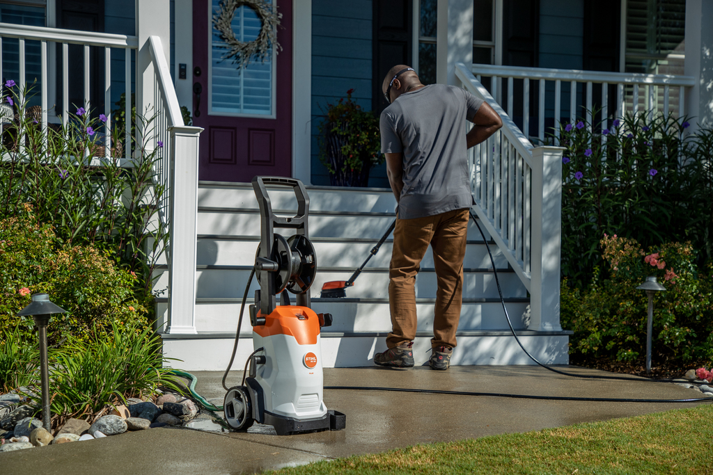 Orange and white STIHL RE 90 Plus pressure washer on driveway with person cleaning concrete steps in a home setting