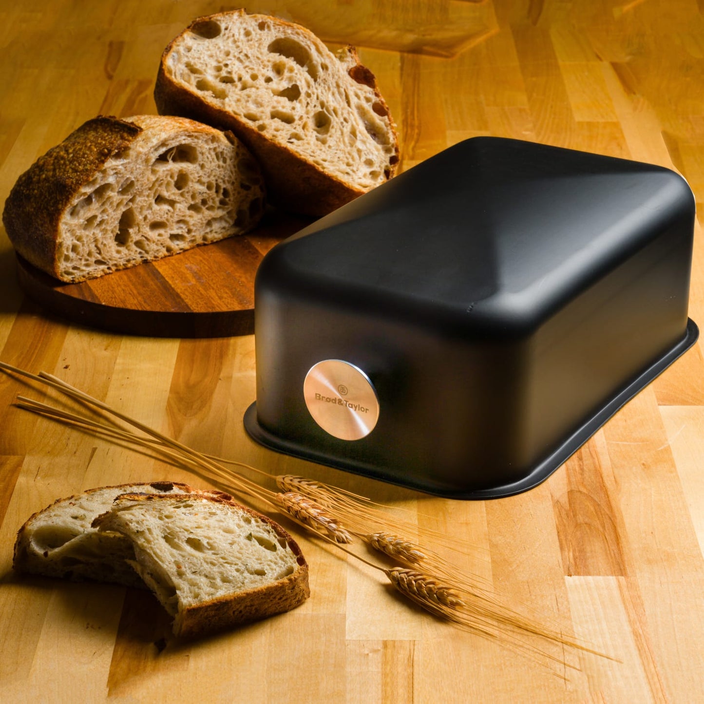 Matte black Brod & Taylor baking shell with rounded edges on wooden surface near sliced artisan bread and wheat stalks