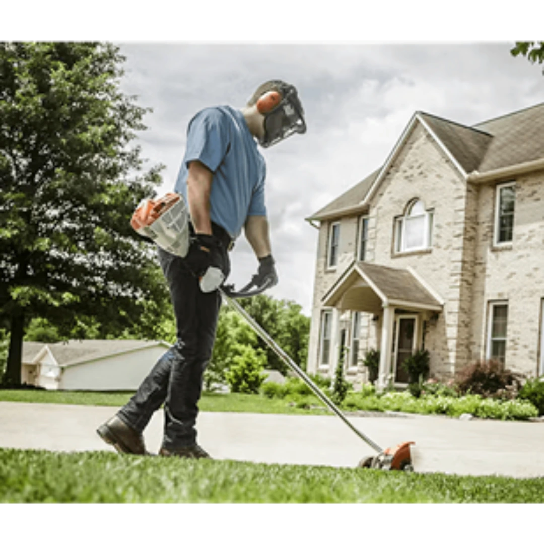 Person wearing protective gear operating a curved shaft edger along a driveway near a brick house for lawn care.