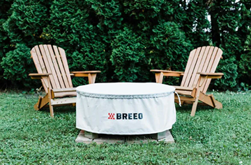 Round white Breeo X19 fire pit cover protecting fire pit outdoors between two wooden chairs on grass