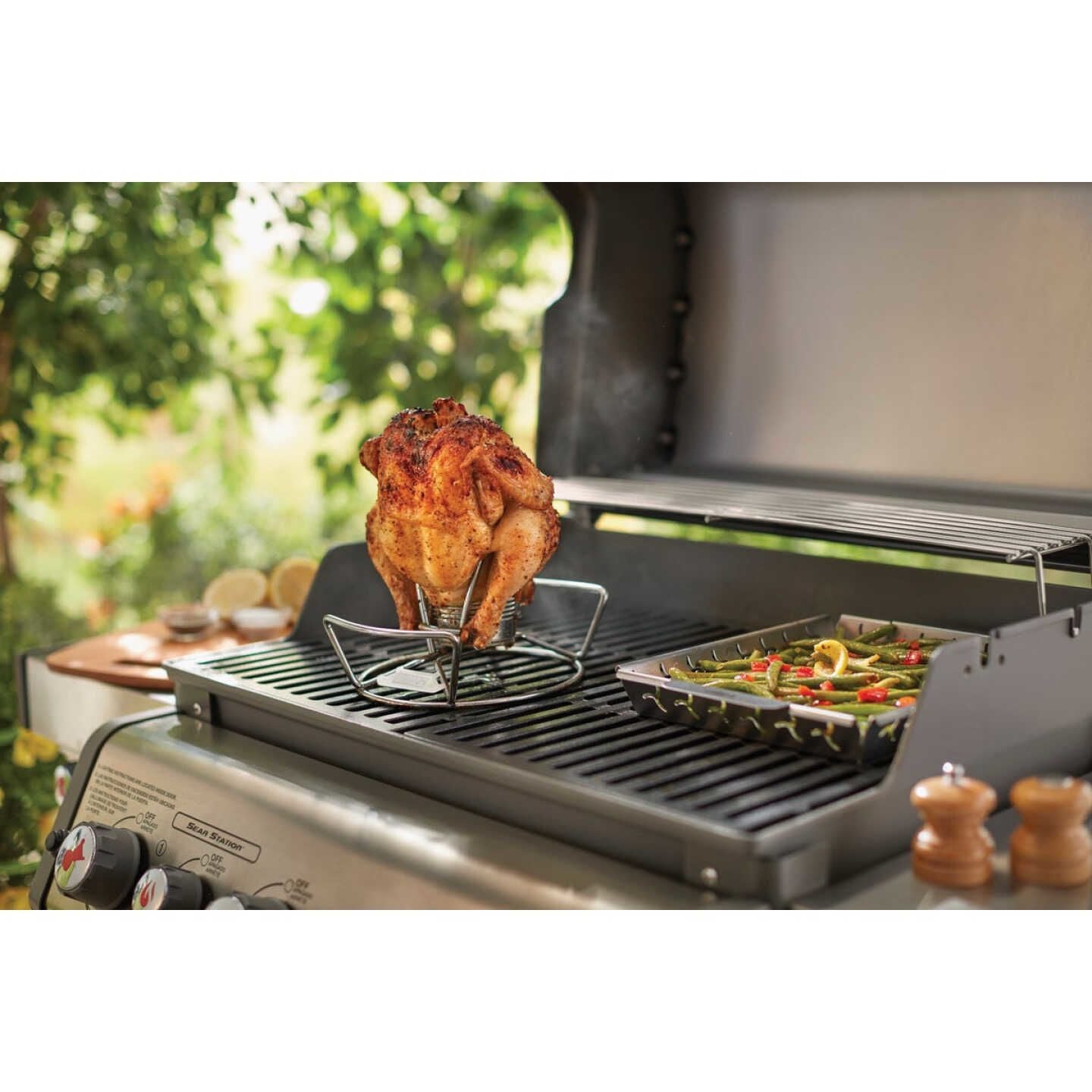 Steel nesting poultry roaster holding seasoned whole chicken upright cooking on gas grill with vegetable tray nearby