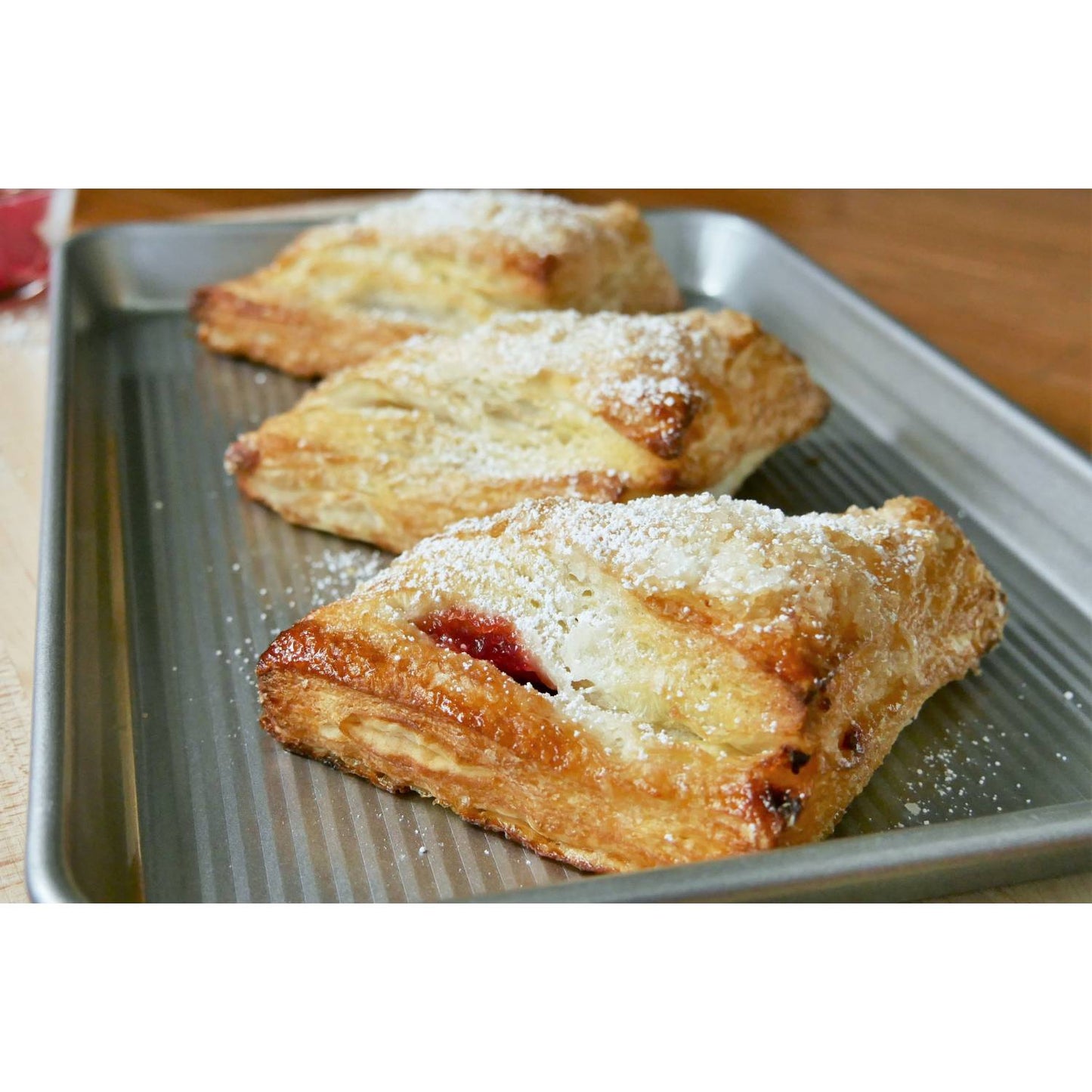 Metal jelly roll pan with baked pastries dusted with powdered sugar on wooden table surface