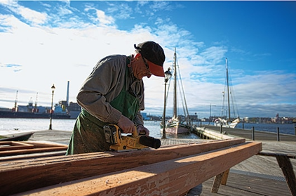 Man using DEWALT cordless brushless planer on wooden beam outdoors near marina for home improvement tasks