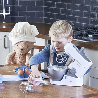 Young boy playing with a Casdon Kenwood Mixer toy in a kitchen setting with a stuffed bear wearing a chef hat nearby