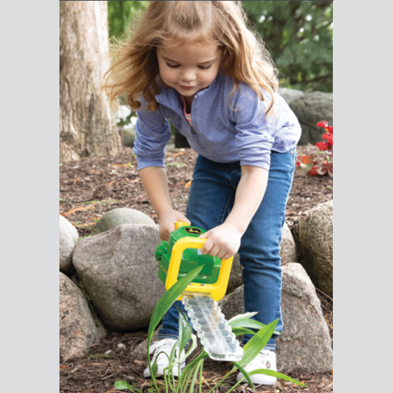 Young child using green and yellow toy hedge trimmer for outdoor play in a garden setting with rocks and plants