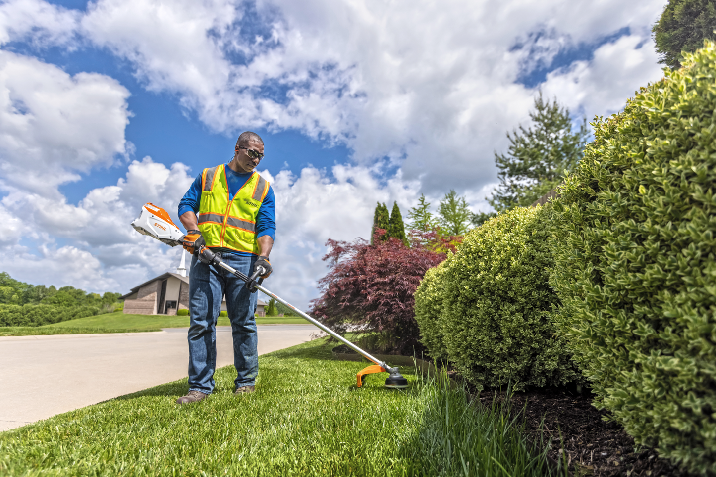 Man wearing safety vest using orange and white battery-powered trimmer to edge grass along garden shrubs outdoors