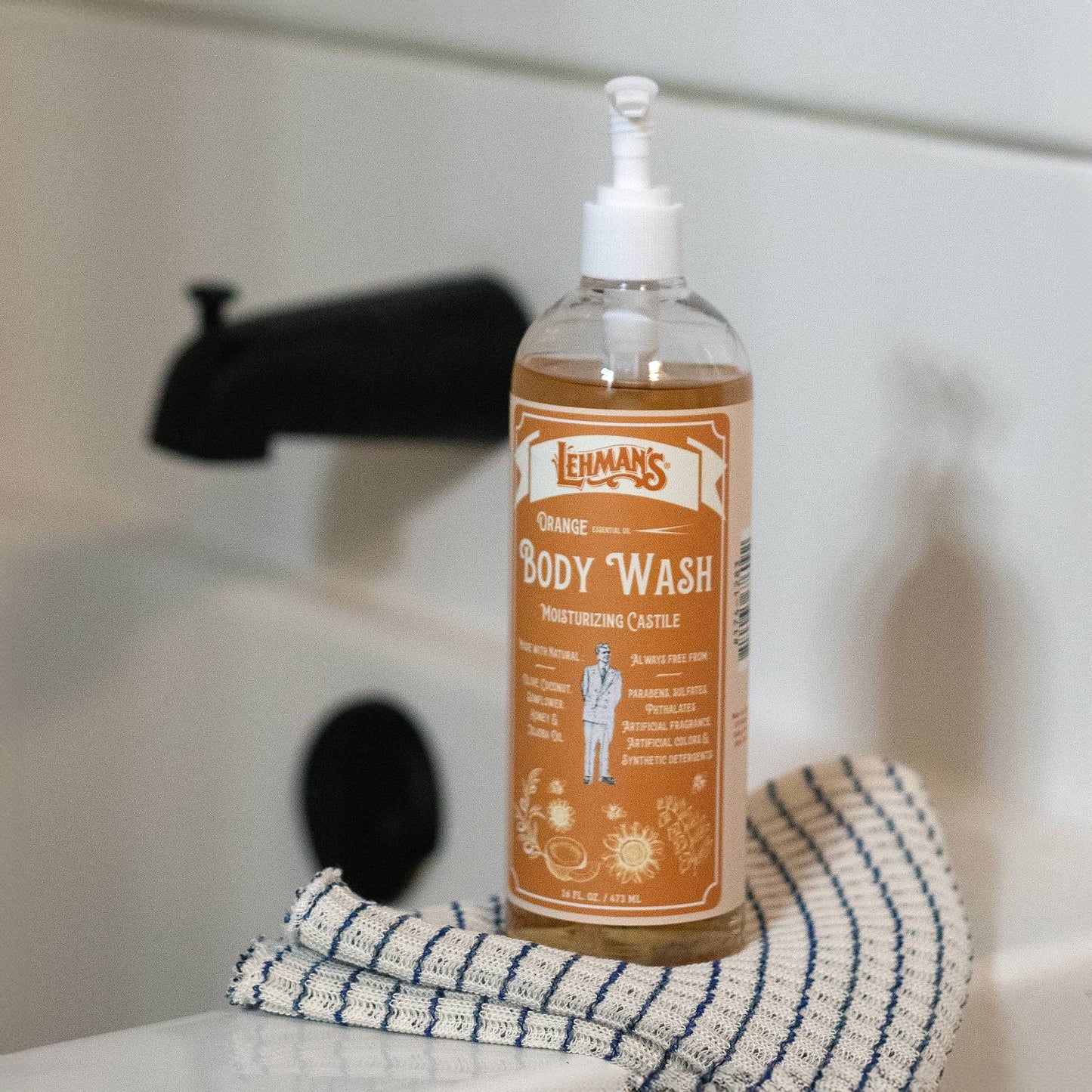 Clear plastic pump bottle of orange-scented moisturizing castile body wash on striped cloth by black faucet, ideal for Health & Beauty and Under $50 collections