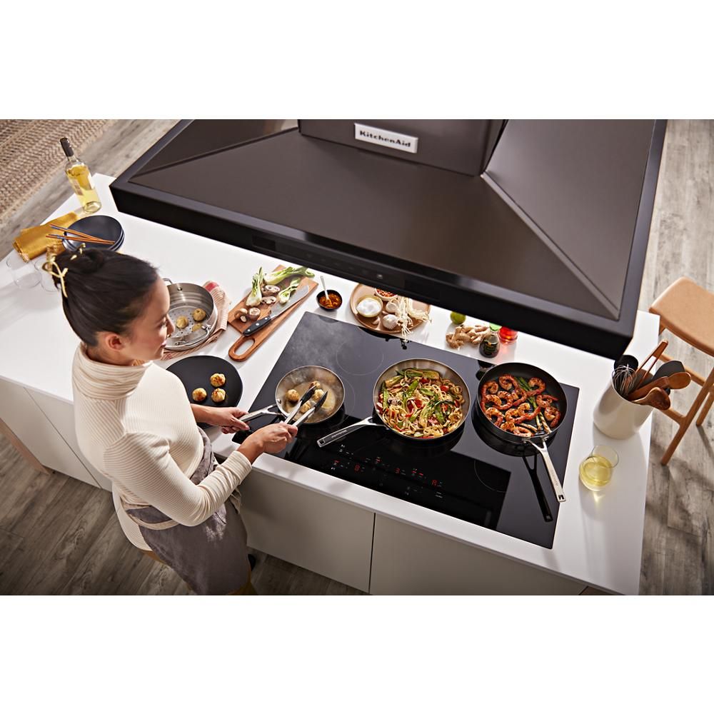 Woman preparing food on black cooktop beneath dark canopy hood in bright kitchen from KitchenAid range hoods collection