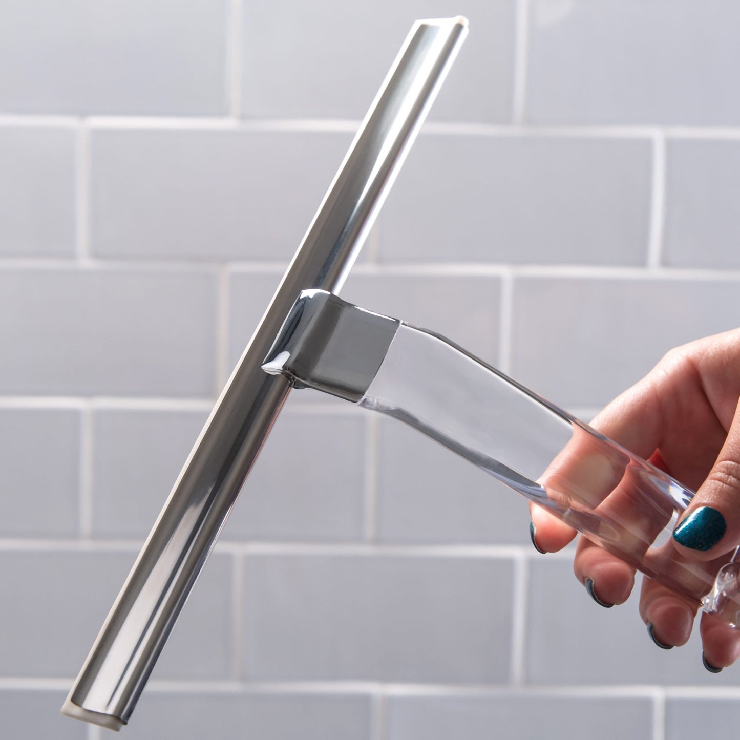 Hand holding clear-handled squeegee with metal blade and hook against a white tiled background for home use