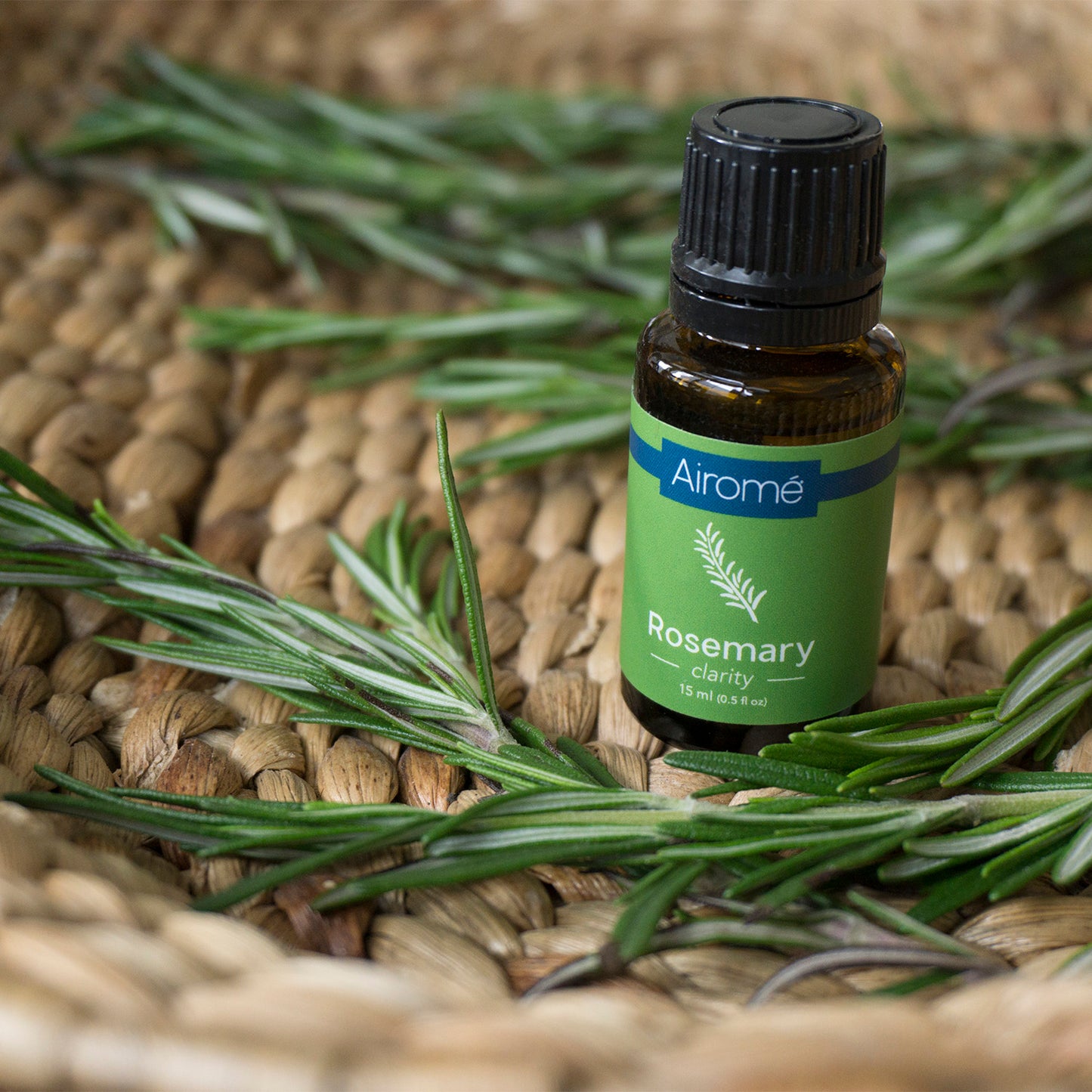Small amber glass bottle of rosemary essential oil surrounded by fresh rosemary sprigs on woven decor surface