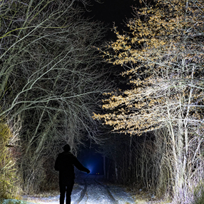 Person shining powerful 15000 lumen tactical flashlight illuminating trees on snowy wooded trail at night