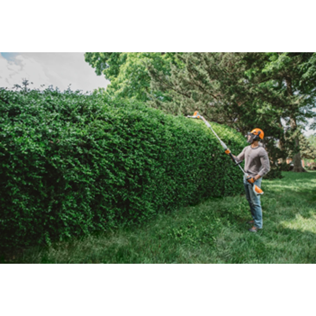 Person using an extended-reach battery hedge trimmer to neatly shape a tall outdoor hedge in a garden setting