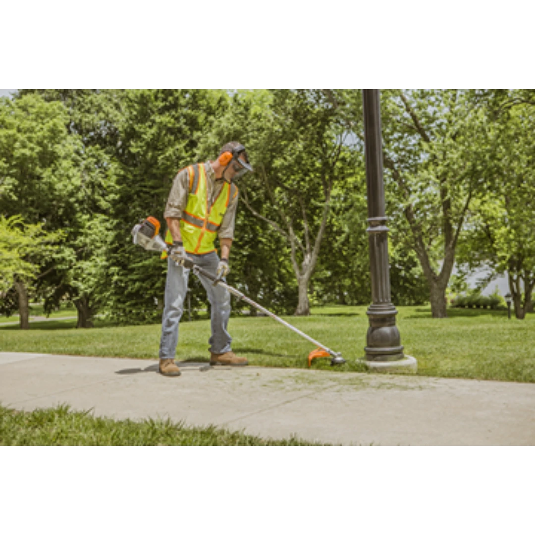 Worker in safety gear using a cordless weed trimmer to edge grass along a sidewalk near a lamppost outdoors