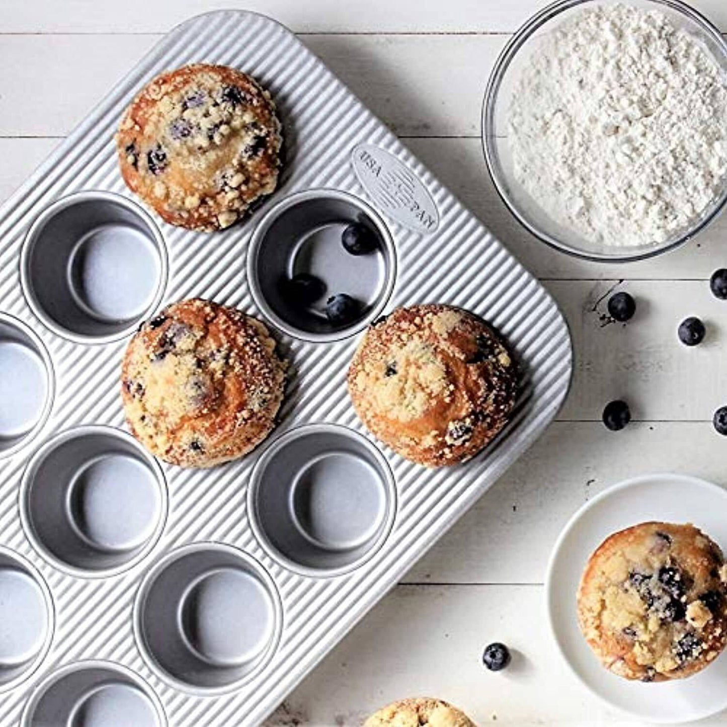 Textured USA PAN metal muffin pan with twelve cups, three baked blueberry muffins, scattered blueberries, and a bowl of flour nearby