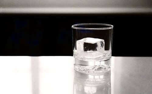 Clear ice cube in a glass tumbler on a reflective surface, suitable for kitchen and bar appliance display