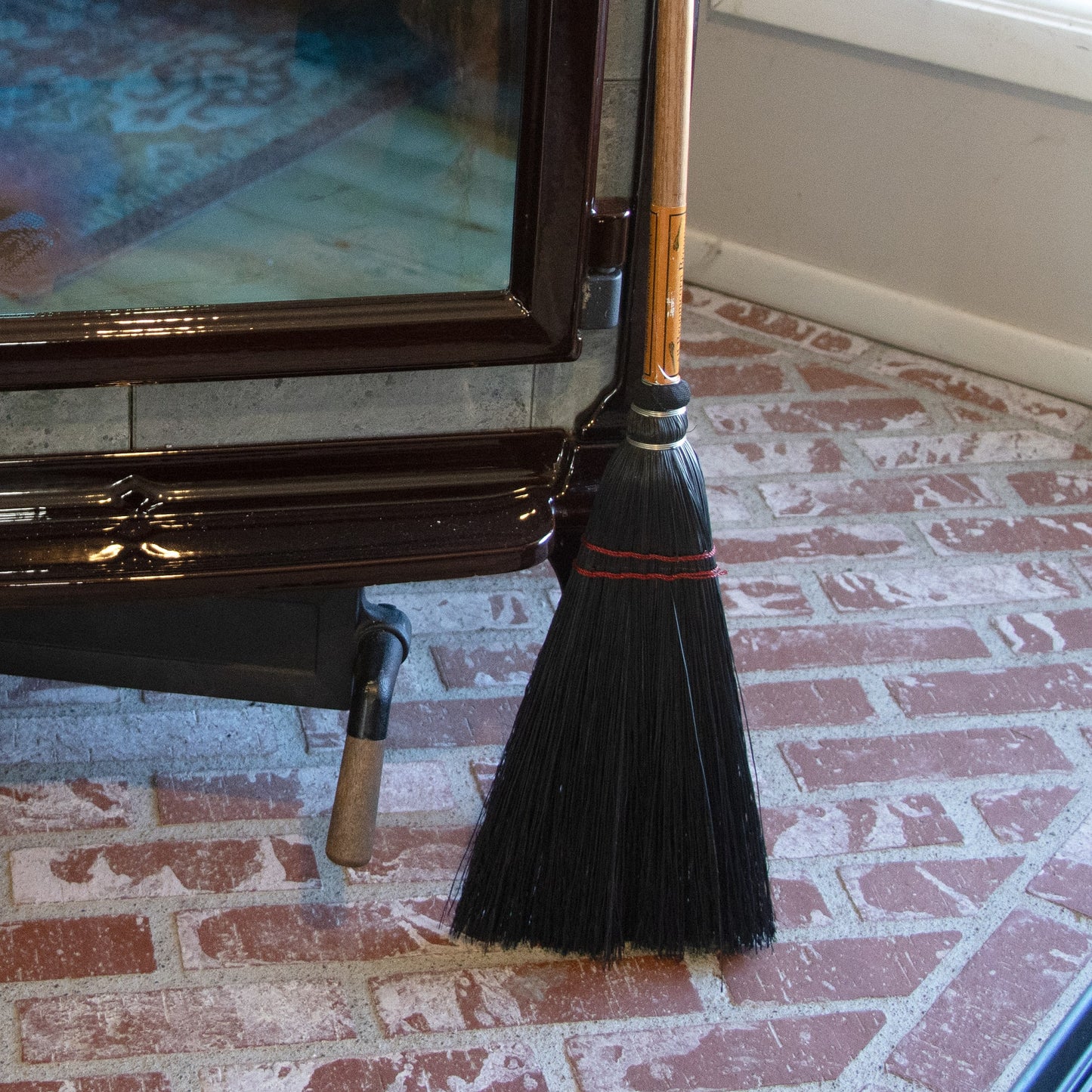 Black bristle broom with wooden handle standing on brick floor next to a black metal stove in cleaning tools collection