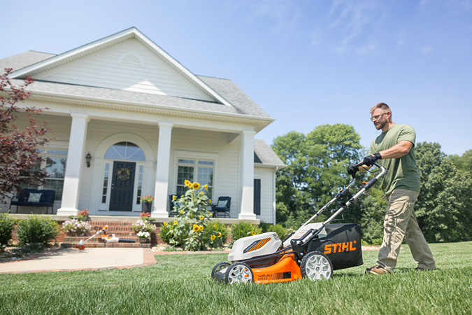Man using an orange and white STIHL battery powered lawn mower with brushless motor and steel deck on a green lawn in front of a white house for outdoor power equipment and home improvement