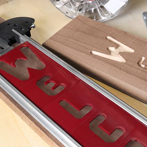 Red vertical letter stencil attached to wood with carved letter W and router bit in woodworking setup