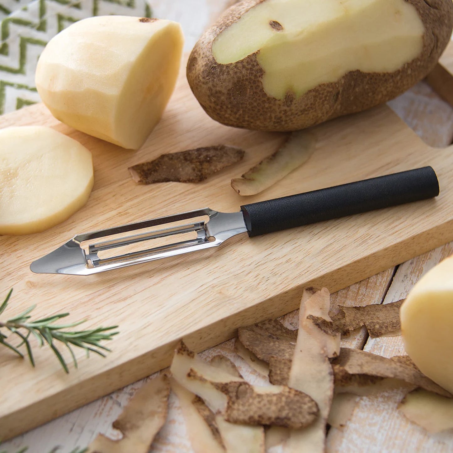 Rada Vegetable Peeler with black resin handle on wooden board with peeled potato, skins, and rosemary in kitchenware setting