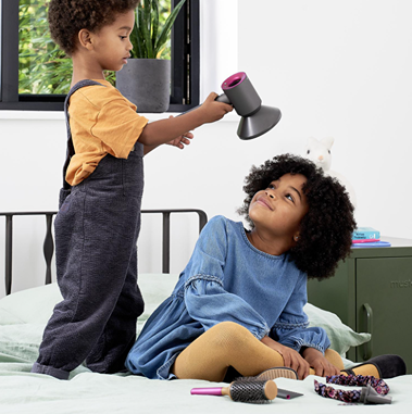 Child playing with a toy hair dryer styling a sibling’s hair in a bright bedroom setting