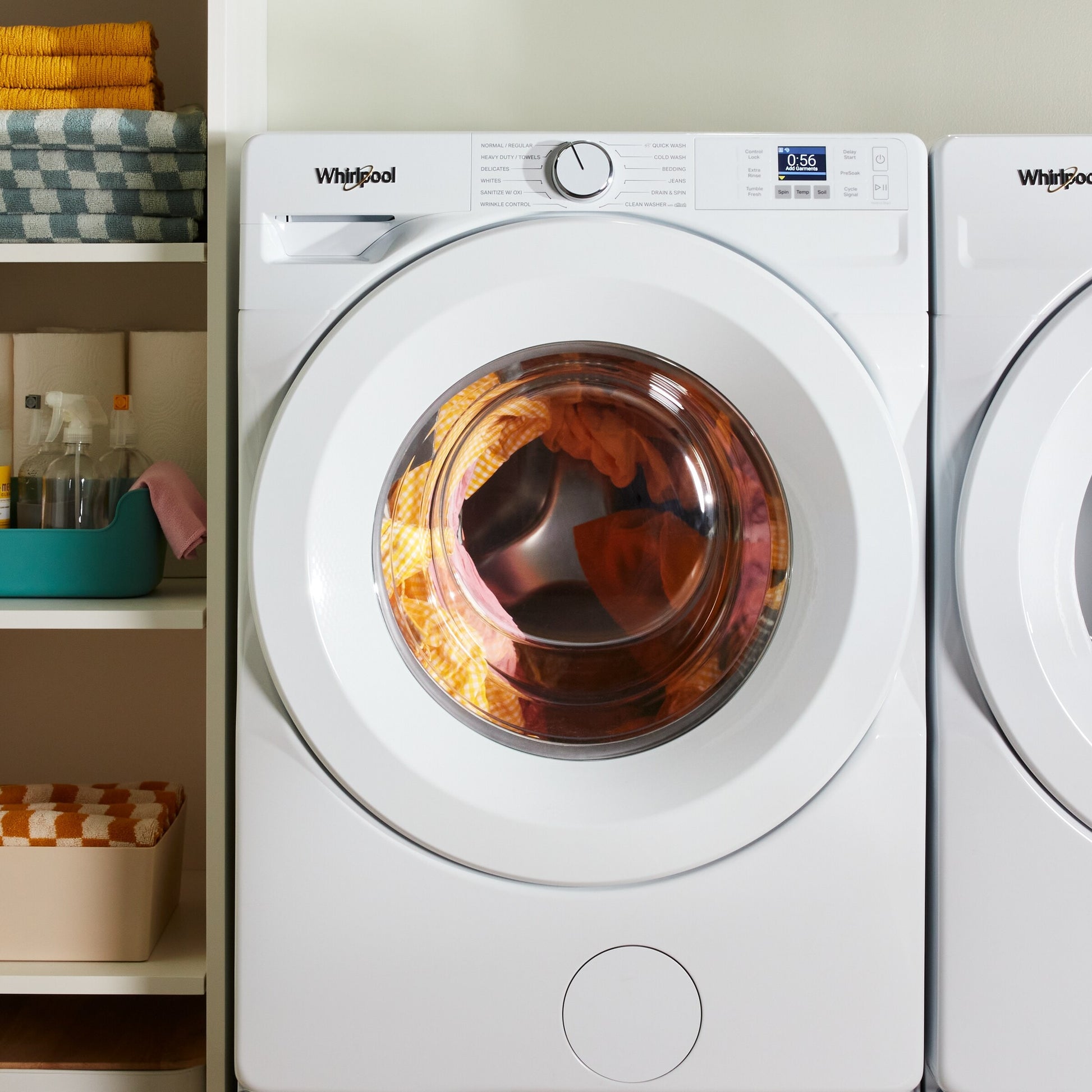 White front load washer with digital display and dial controls, shown in laundry room with shelves and folded towels nearby