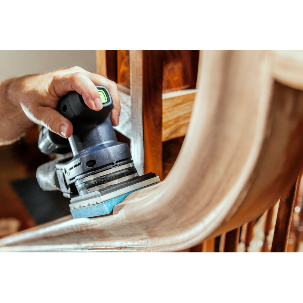 Person using handheld power sander with blue foam-backed abrasive on curved wooden stair railing in home improvement setting