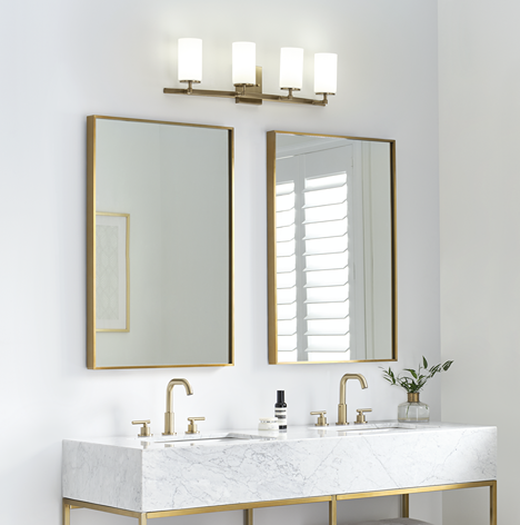 Four-light satin brass wall fixture with cylindrical etched glass shades above double bathroom vanity mirrors