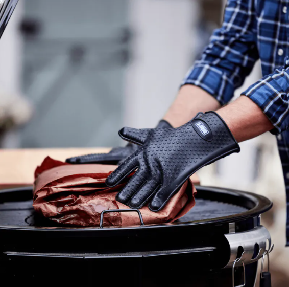 Black silicone grilling gloves with textured grip handling a wrapped item on a charcoal grill in outdoor cooking setting